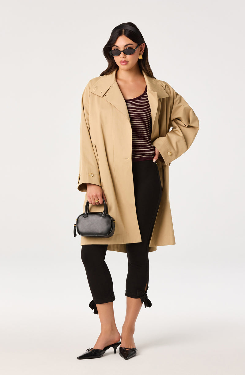 Woman wearing a beige trench coat, black capri pants with bow details, black pointed shoes, and carrying a small black handbag.