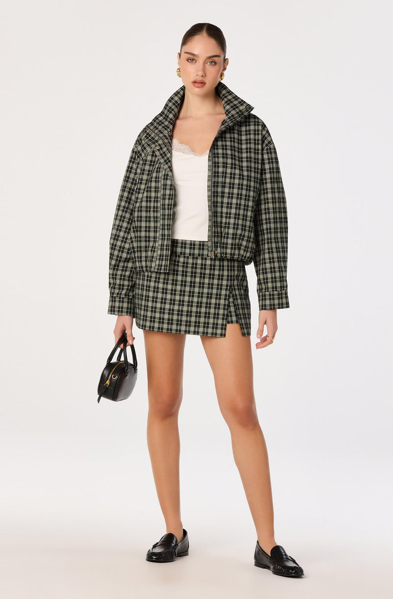 Model wears a green and black plaid jacket, matching plaid mini skirt, white lace-trimmed blouse, black loafers, and gold earrings, holding a small black handbag.