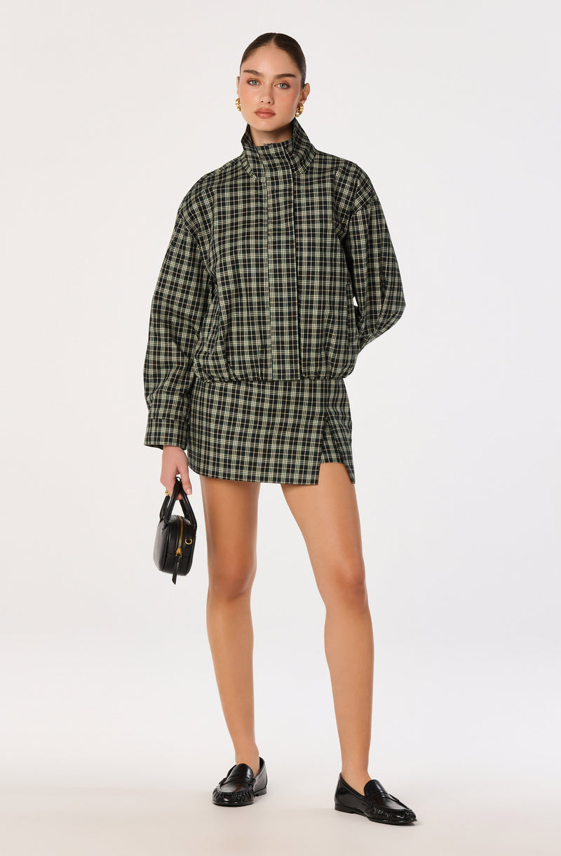 Model wearing a black and green plaid jacket and matching skirt with high boots.