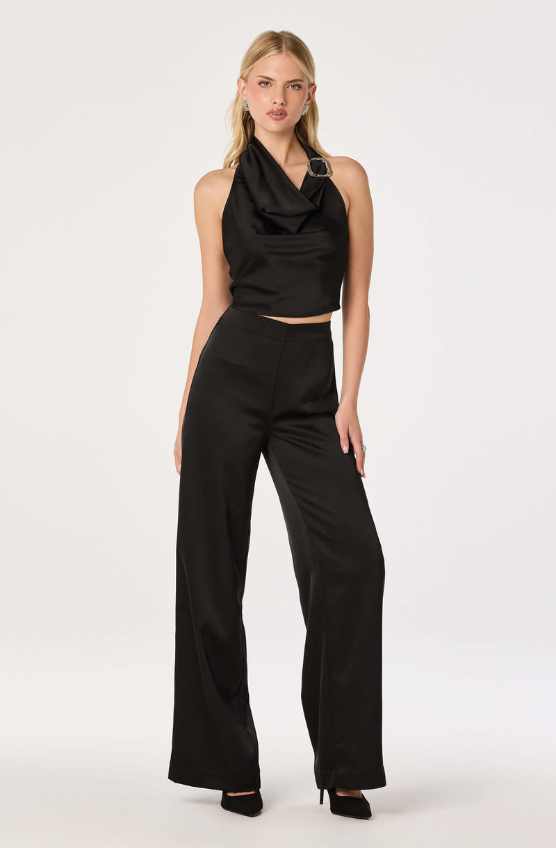 Woman wearing a sleeveless black satin top with a draped neckline and a buckle detail, paired with high-waisted black wide-leg pants.