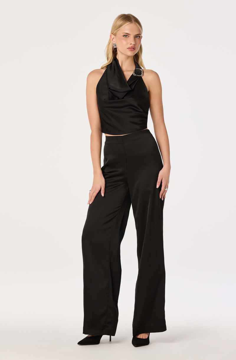 Woman wearing a black sleeveless top with a draped collar and large buckle detail, paired with high-waisted black wide-leg pants and black heels.