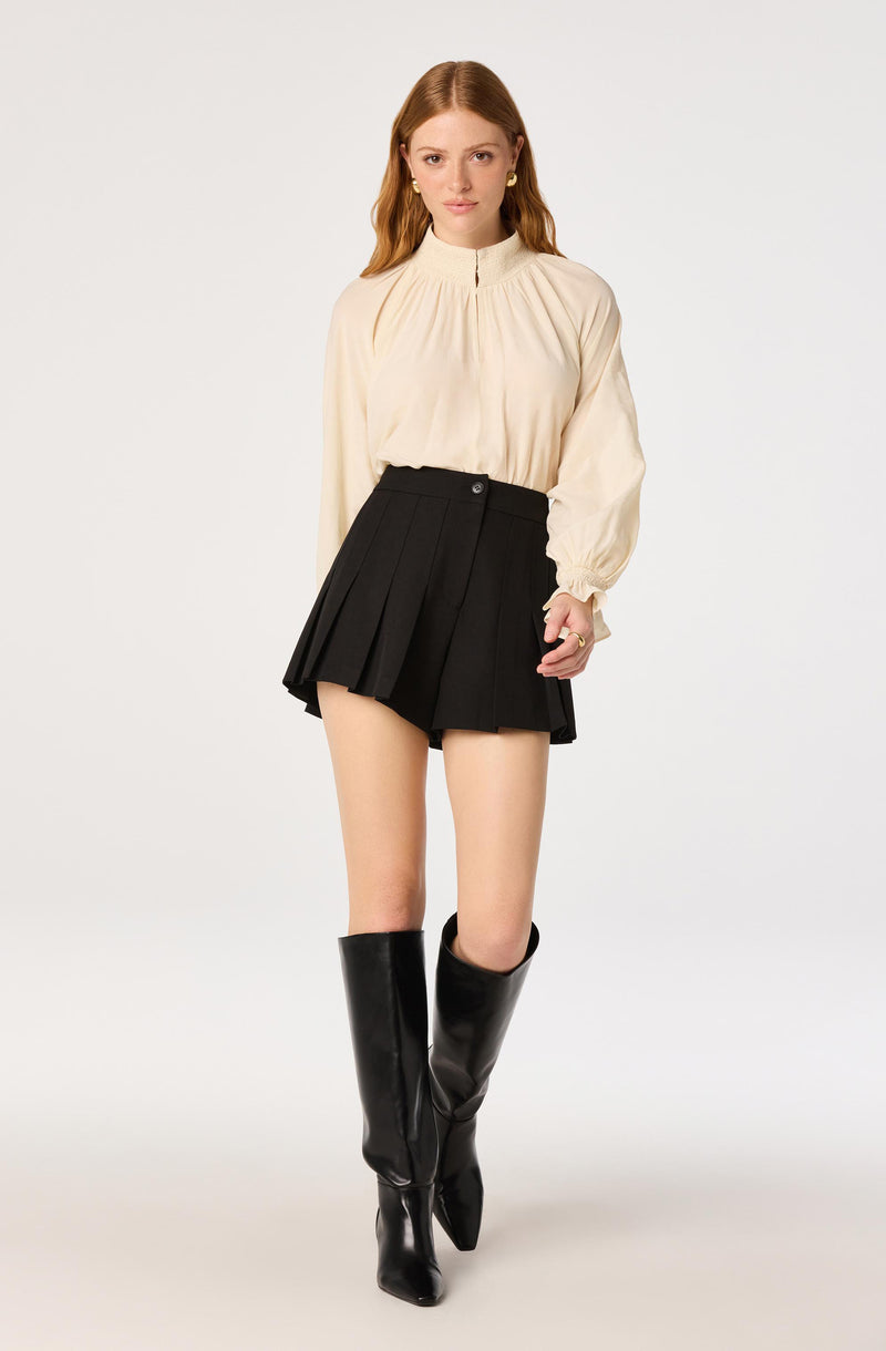 Woman wearing a cream blouse, black pleated shorts, and knee-high black boots.