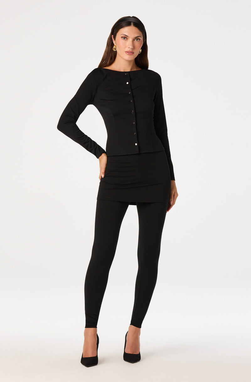 Woman in all-black outfit with a fitted long-sleeve top, matching skirt, leggings, and black high heels.