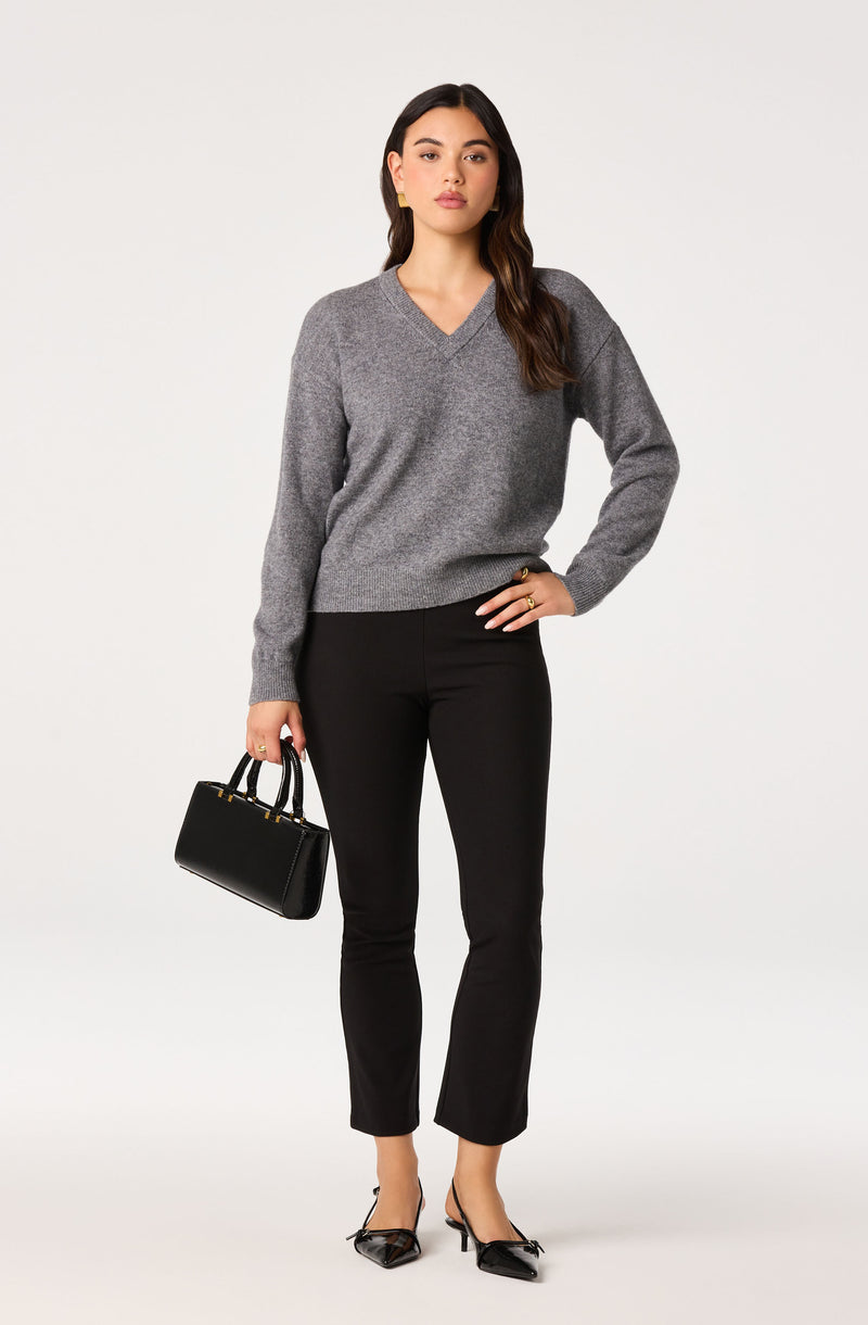 Woman in a gray V-neck sweater, black pants, black heels, holding a black handbag.