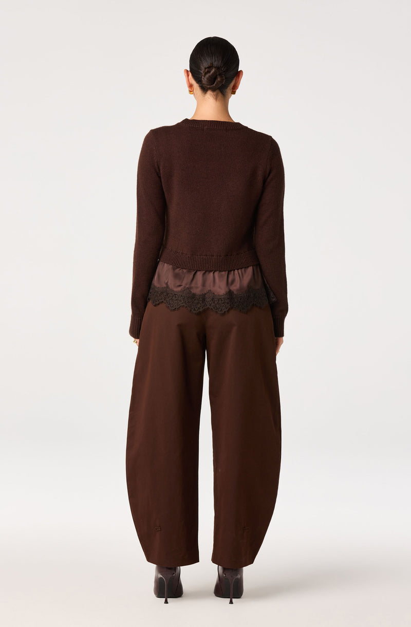 Woman wearing a dark brown sweater with a ruffled, satin and lace trim at the hem, and loose brown wide-leg pants.