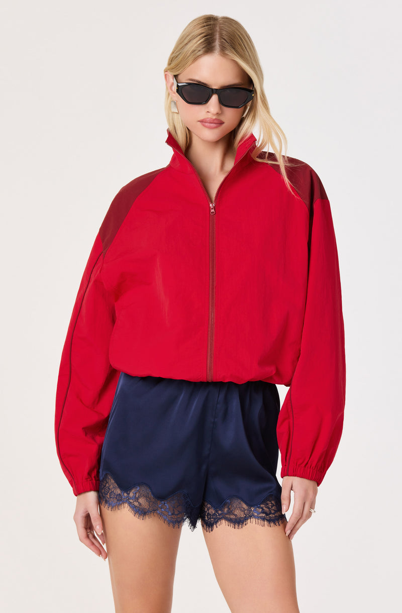 Woman in a red and black athletic jacket, navy silk shorts with lace trim, wearing black sunglasses.