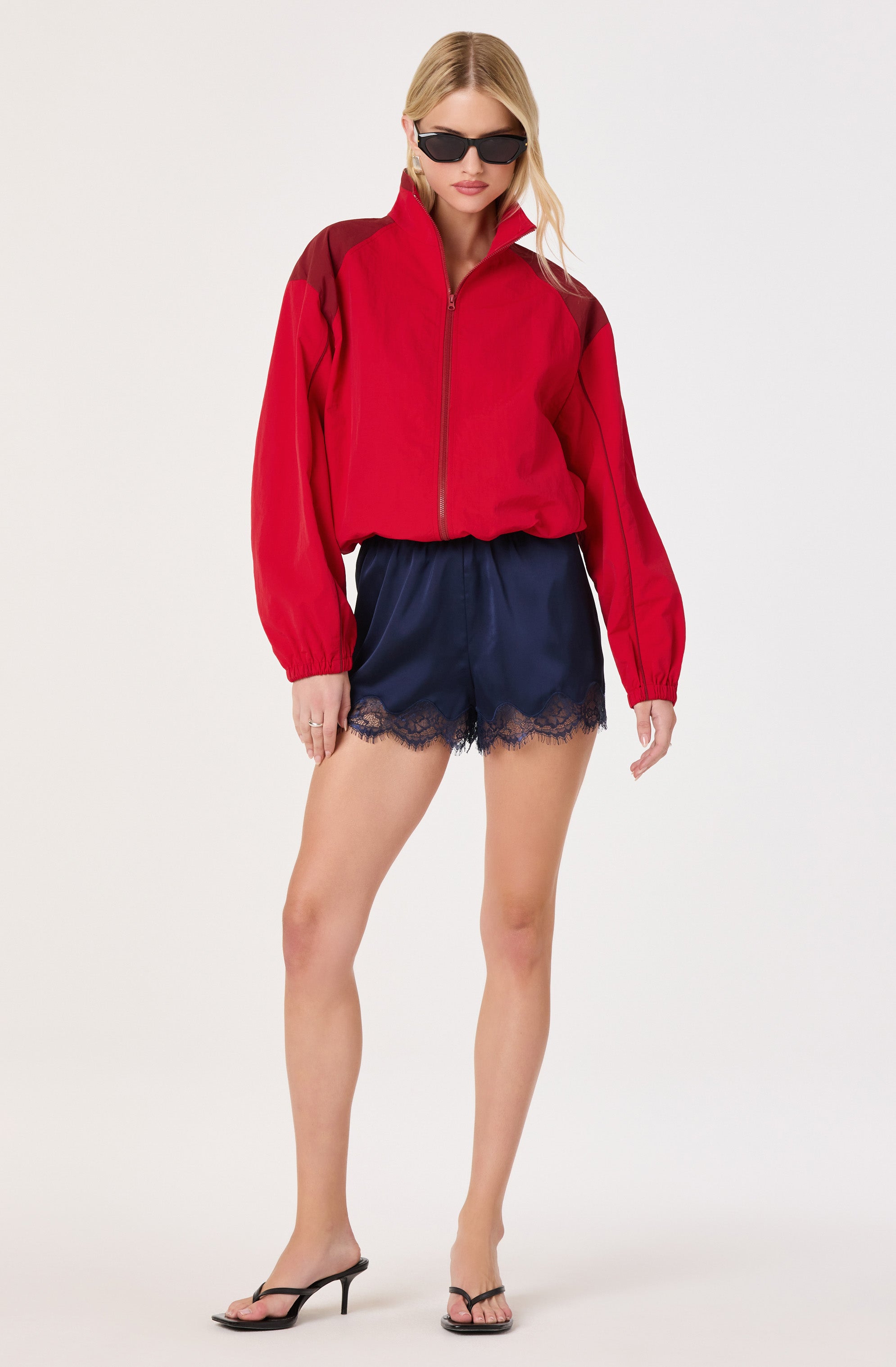 Woman in a red and black zip-up jacket, navy satin shorts with lace trim, black high heels, and sunglasses.
