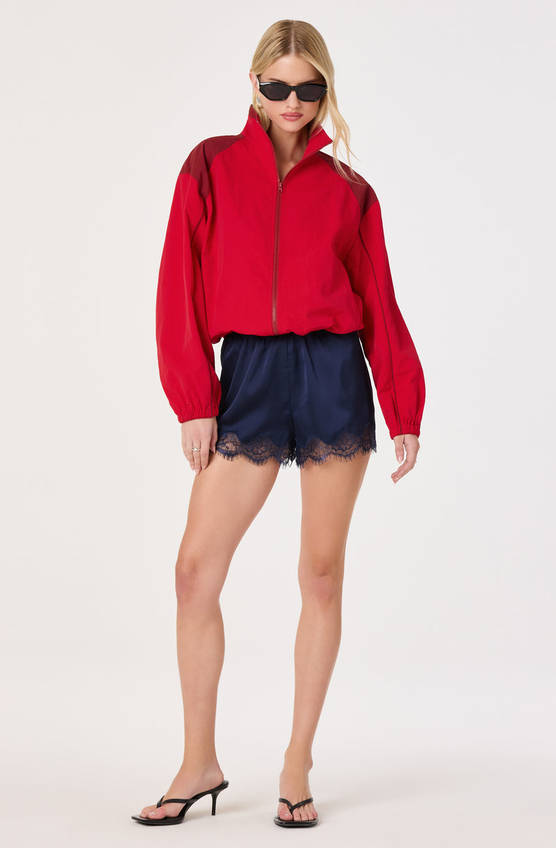 Woman in a red and black zip-up jacket, navy satin shorts with lace trim, black high heels, and sunglasses.