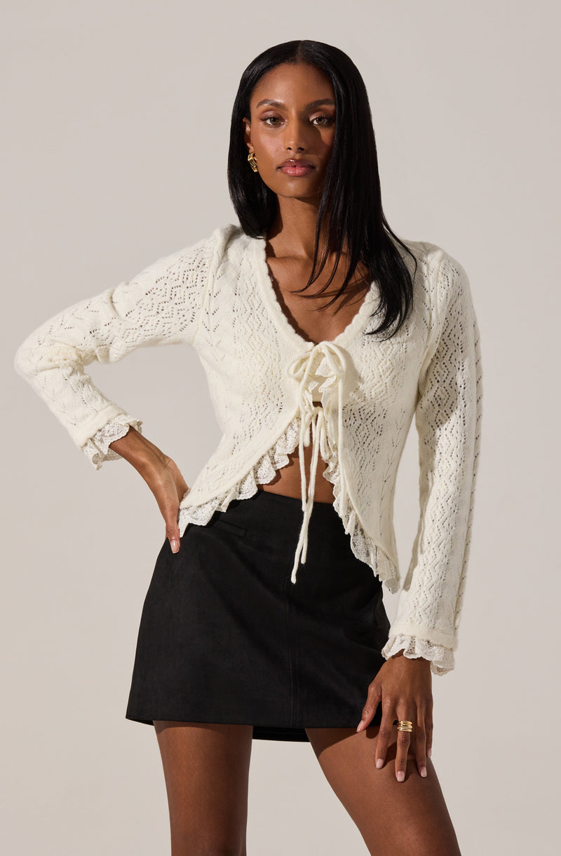 A woman is wearing a cream-colored, long-sleeved, knitted cardigan with a lace pattern. The cardigan features a ruffled hem and cuffs, and it is tied at the front with a bow. She pairs it with a black, high-waisted, A-line skirt.