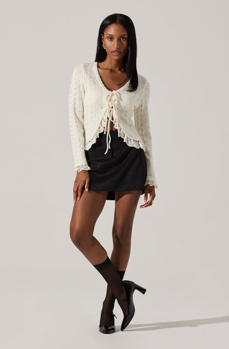 A woman is wearing a cream-colored, long-sleeved, knitted cardigan with a ruffled hem and a bow tie at the front. She pairs it with a black mini skirt, sheer black ankle socks, and black high-heeled shoes.
