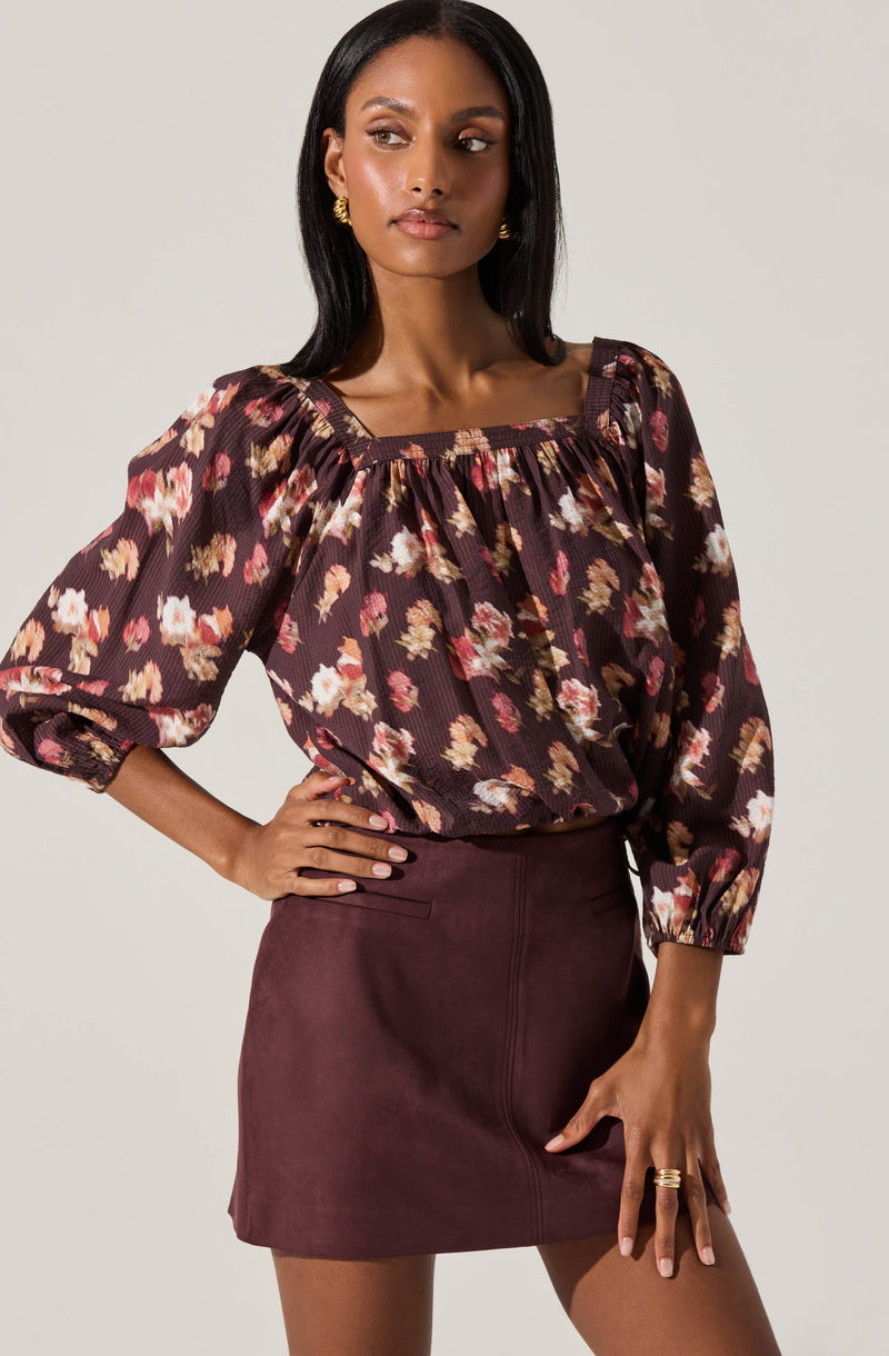A woman is wearing a long-sleeved blouse with a floral pattern featuring shades of pink, beige, and white on a dark background. The blouse has a gathered neckline and puffed sleeves that are elasticized at the cuffs. She pairs it with a high-waisted, maroon-colored skirt that appears to be made of a suede-like material. The skirt has a fitted silhouette with a small slit at the front. The woman accessorizes with gold earrings and rings on her fingers.