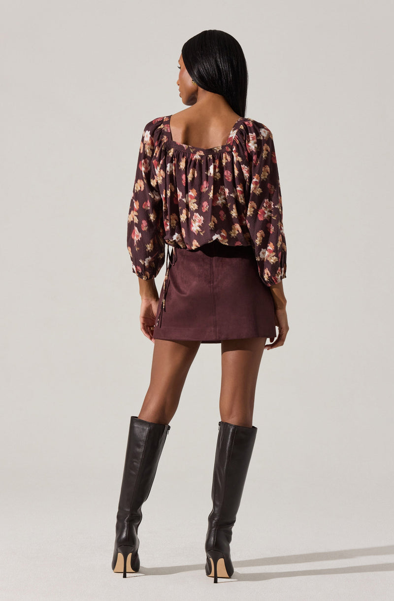 A woman is wearing a long-sleeved, dark-colored blouse with a floral print in shades of pink, peach, and cream. The blouse has a square neckline with gathered fabric detail. She pairs it with a high-waisted, short, burgundy skirt made of corduroy material. She completes her outfit with tall, black leather knee-high boots that have stiletto heels.