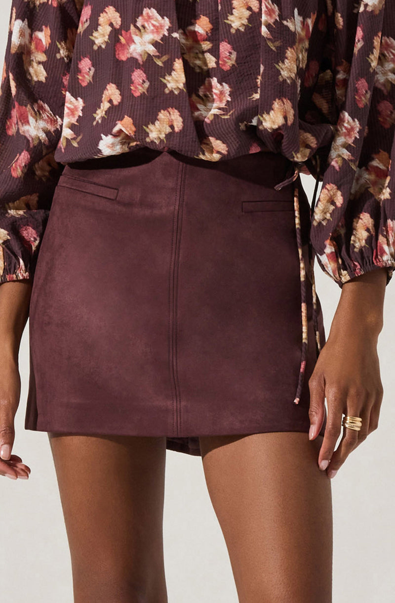 A person is wearing a dark floral blouse with long, puffed sleeves and a drawstring at the waist, paired with a high-waisted, burgundy mini skirt made of a suede-like material.