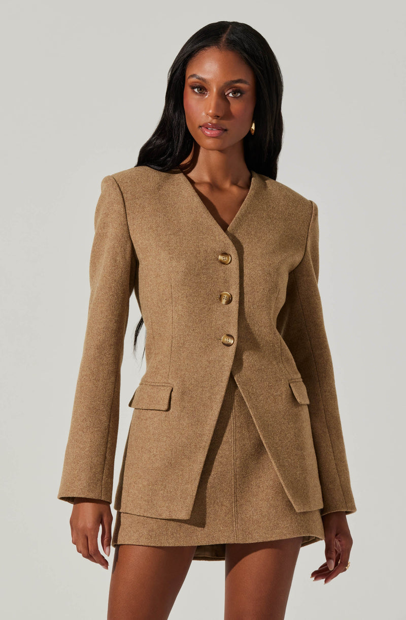 A woman is wearing a tailored beige blazer with a structured fit, featuring a V-neckline, long sleeves, and four gold buttons down the front. The blazer has flap pockets on each side at hip level. She pairs it with a matching beige skirt made of the same fabric, which is fitted and ends above the knee.