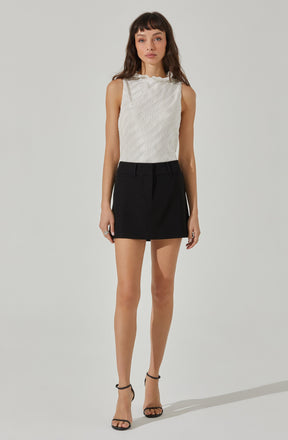 A woman is wearing a sleeveless, light-colored top with a subtle vertical stripe pattern and ruffled neckline paired with a high-waisted black mini skirt. She has on black high-heeled sandals with ankle straps.