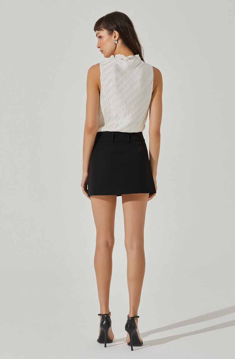 A woman is wearing a sleeveless, high-neck, white textured top with subtle vertical stripes. She pairs it with a black mini skirt that has a clean, straight cut and belt loops. She completes the outfit with black high-heeled shoes that have ankle straps.