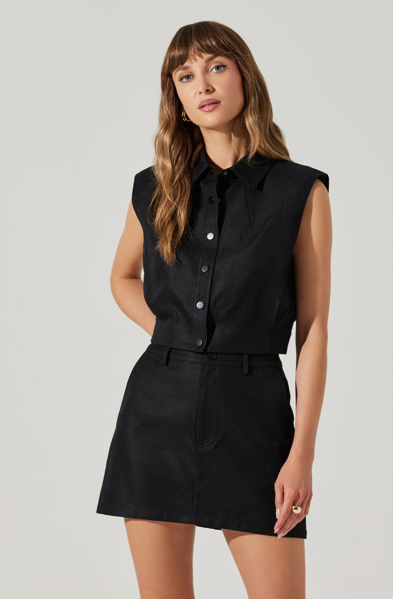 A woman is wearing a sleeveless black button-up shirt with a pointed collar and padded shoulders, paired with a high-waisted, black denim mini skirt.