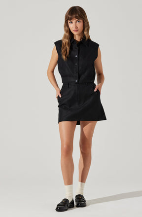 A woman is wearing a sleeveless black denim shirt with a collar and buttons down the front. She pairs it with a high-waisted black skirt that has a slight A-line shape and belt loops. She completes her outfit with white crew socks and black slip-on shoes with buckle detail.