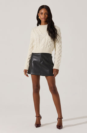 A woman is wearing a cream-colored, cable-knit sweater with a high neckline, paired with a black leather mini skirt. She has on dark brown high heels with ankle straps and gold jewelry, including earrings and rings.