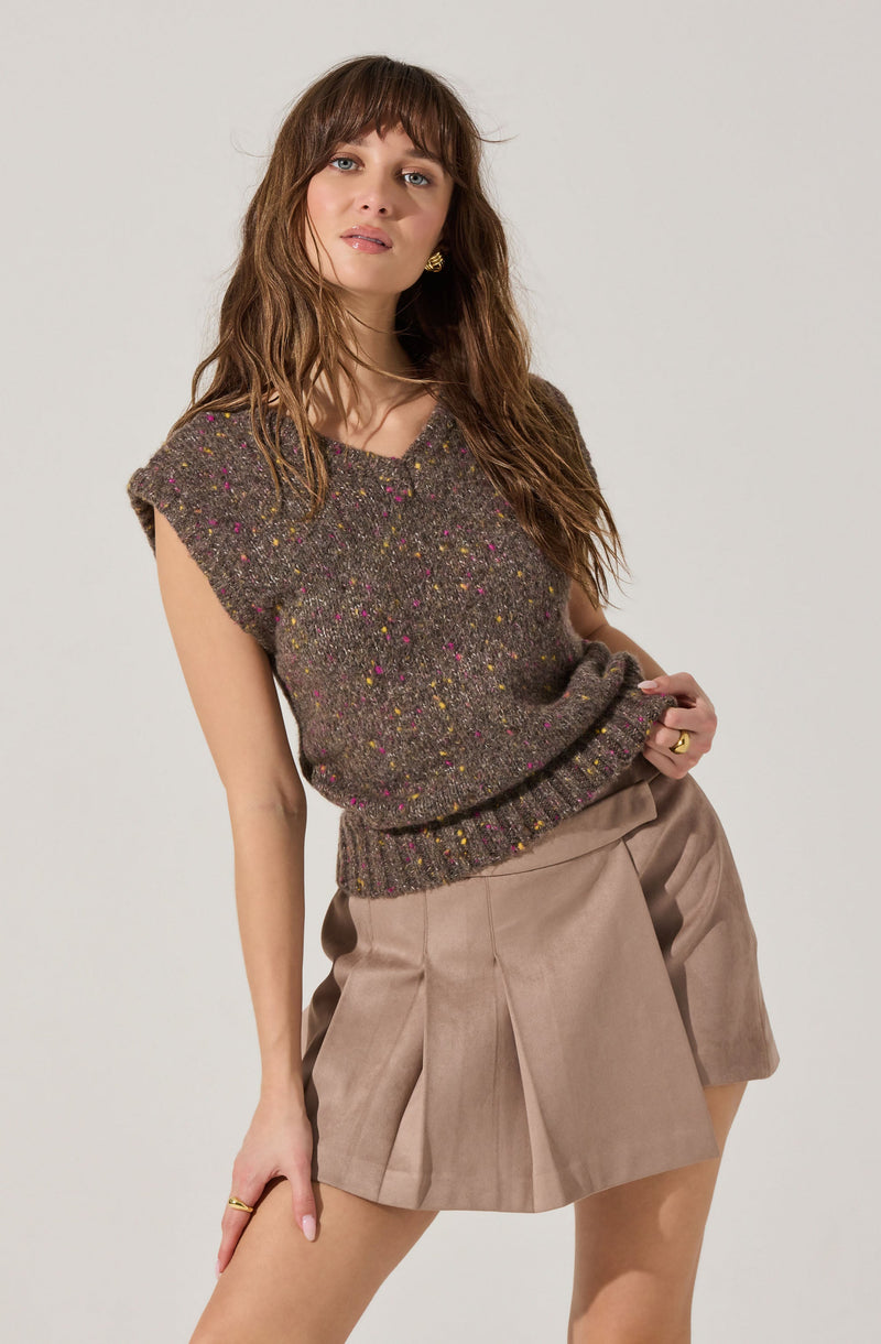 A woman is wearing a sleeveless, knit sweater in a dark brown color with multicolored specks throughout. The sweater has a V-neckline and a ribbed hem. She pairs it with beige pleated shorts.