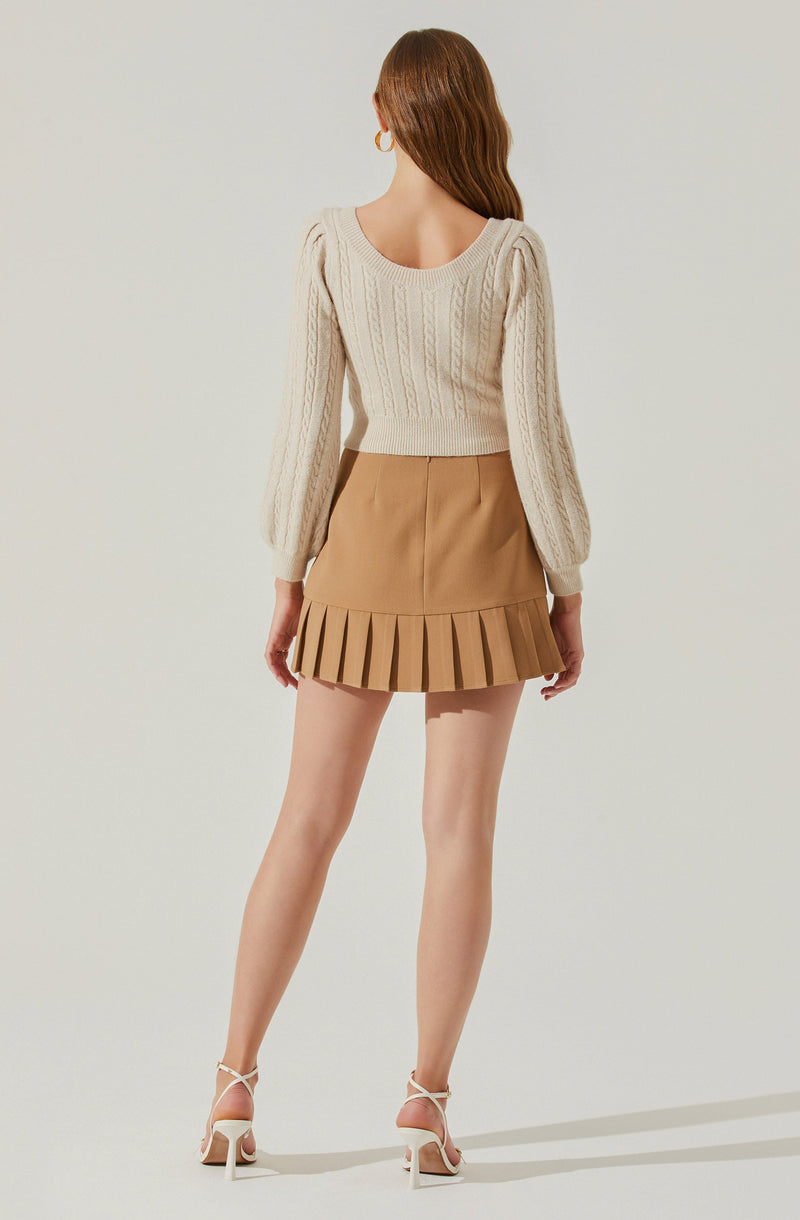 A woman is wearing a cream-colored, long-sleeve, cable-knit sweater with a scoop neckline, paired with a tan mini skirt that features a pleated hem. She has on white high-heeled sandals with ankle straps.