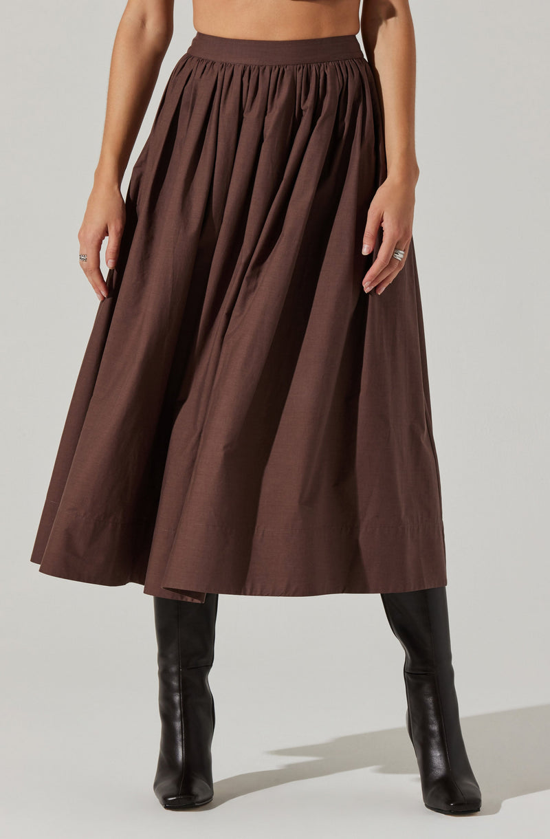 A woman is wearing a high-waisted, full-length brown skirt with pleats, paired with tall black boots.