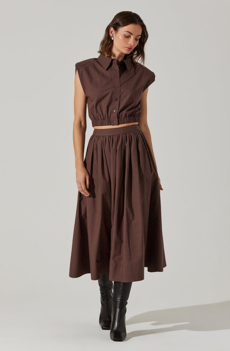 A woman is wearing a sleeveless, button-up brown top with shoulder pads and an elastic waistband, paired with a matching high-waisted, full-length brown skirt with pleats. She completes her outfit with tall black boots.