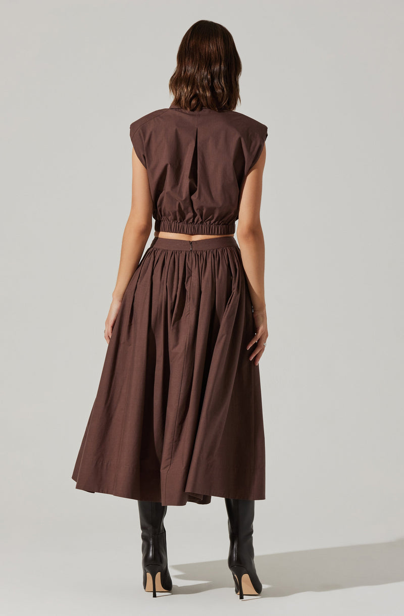 The outfit consists of a brown sleeveless cropped top with shoulder padding and an elastic waistband, paired with a matching high-waisted, full-length skirt that has gathered fabric and pleats. The look is completed with knee-high black heeled boots.