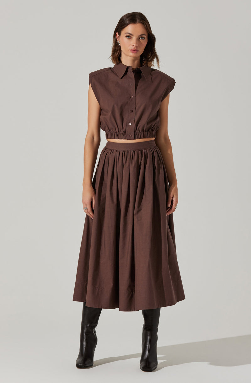 A woman is wearing a sleeveless, cropped, brown button-up shirt with a collar and an elastic waistband. She is also wearing a matching high-waisted, full-length, pleated brown skirt and black knee-high boots.