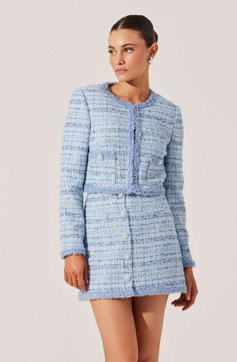 A woman is wearing a matching light blue tweed jacket and skirt set. The jacket is cropped with long sleeves, a round neckline, and decorative buttons on the front and on the sides. The skirt is high-waisted with a row of buttons down the front. The fabric has a textured, woven appearance with a mix of light blue, white, and darker blue threads. The woman has dark hair pulled back and is accessorized with hoop earrings.
