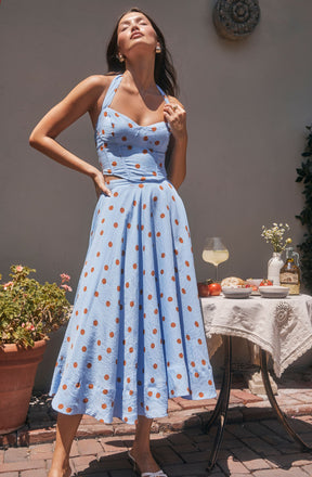 A woman is wearing a light blue sleeveless dress with wide straps and a fitted bodice. The dress features a pattern of evenly spaced brown polka dots. The skirt of the dress is full and long, reaching down to her ankles.