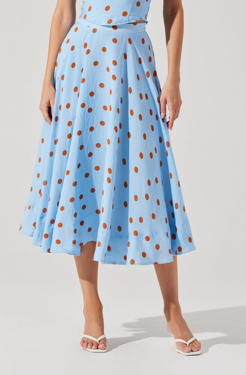 A woman is wearing a light blue, ankle-length skirt with an orange polka dot pattern. The skirt has a high waist and pleats, creating a full, flowing silhouette. She is also wearing a matching light blue top with the same orange polka dots.