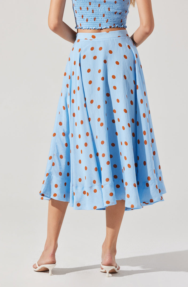 A woman is wearing a high-waisted, light blue midi skirt with an orange polka dot pattern. The skirt has a full, flowing silhouette with pleats, and she is pairing it with a matching sleeveless blue top with a similar orange polka dot pattern. She is also wearing white heeled sandals.