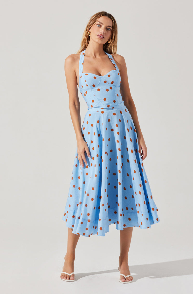 A woman is wearing a sleeveless, light blue dress with a fitted bodice and a sweetheart neckline. The dress features a pattern of evenly spaced brown polka dots and has wide shoulder straps. The skirt is full and midi-length, flowing outward with a pleated design that adds volume and movement. She is also wearing white high-heeled sandals and gold earrings.