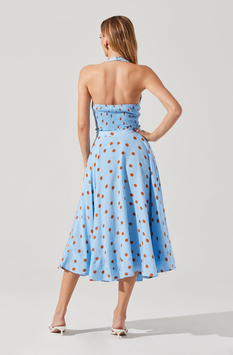 A woman is wearing a strapless light blue dress with orange polka dots. The dress has a fitted, smocked bodice and a full, flowing skirt that reaches mid-calf. She is also wearing white heeled shoes.