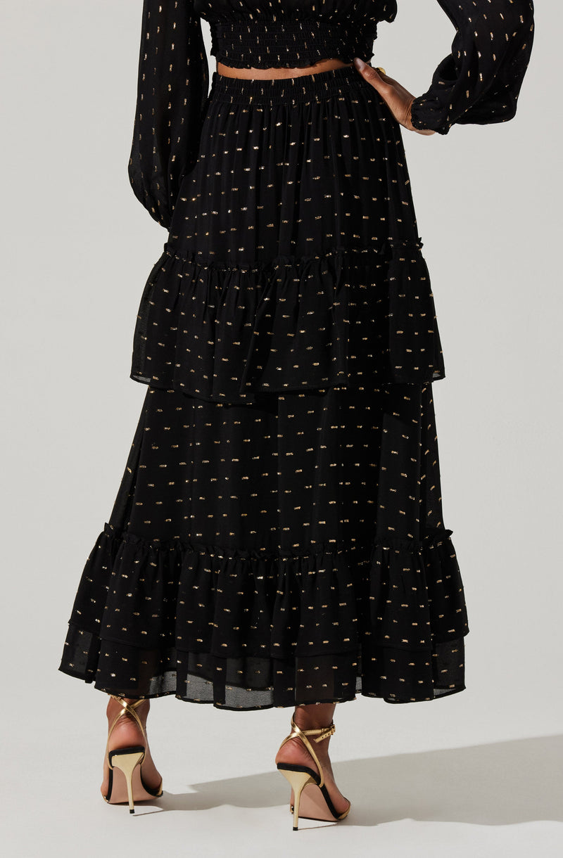 A woman is wearing a black, long, tiered skirt with small, evenly spaced gold dots. The skirt has ruffled tiers at the bottom. She is also wearing a matching black long-sleeved blouse with similar small gold dot patterns, with the sleeves slightly puffed.