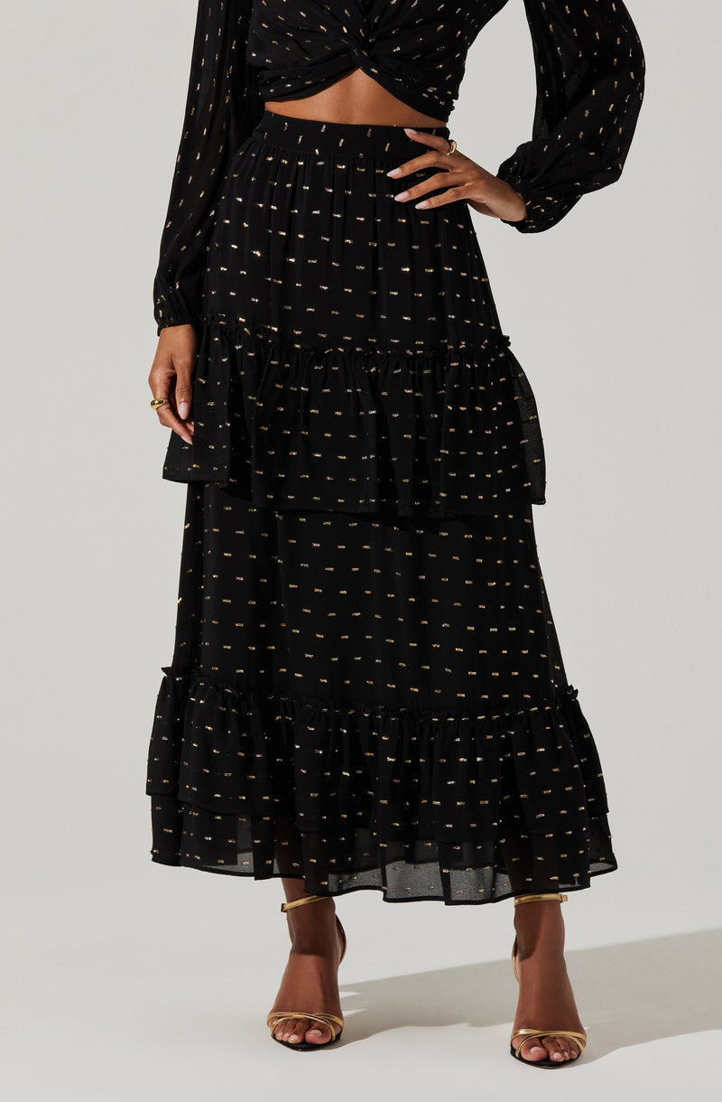 A woman is wearing a long black skirt with gold dotted patterns and ruffled tiered sections. She is also wearing a matching black top with gold dotted patterns, which is tied in a knot at the waist, revealing her midriff. The outfit is complemented by gold high-heeled sandals.