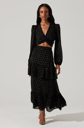 A woman is wearing a black dress with a gold dotted pattern. The dress features long, puffed sleeves and a deep V neckline. The top is twisted at the waist, creating a cutout detail exposing her midriff. The skirt is long and flowing, with layered ruffles that add volume and movement. She is also wearing gold high-heeled sandals.