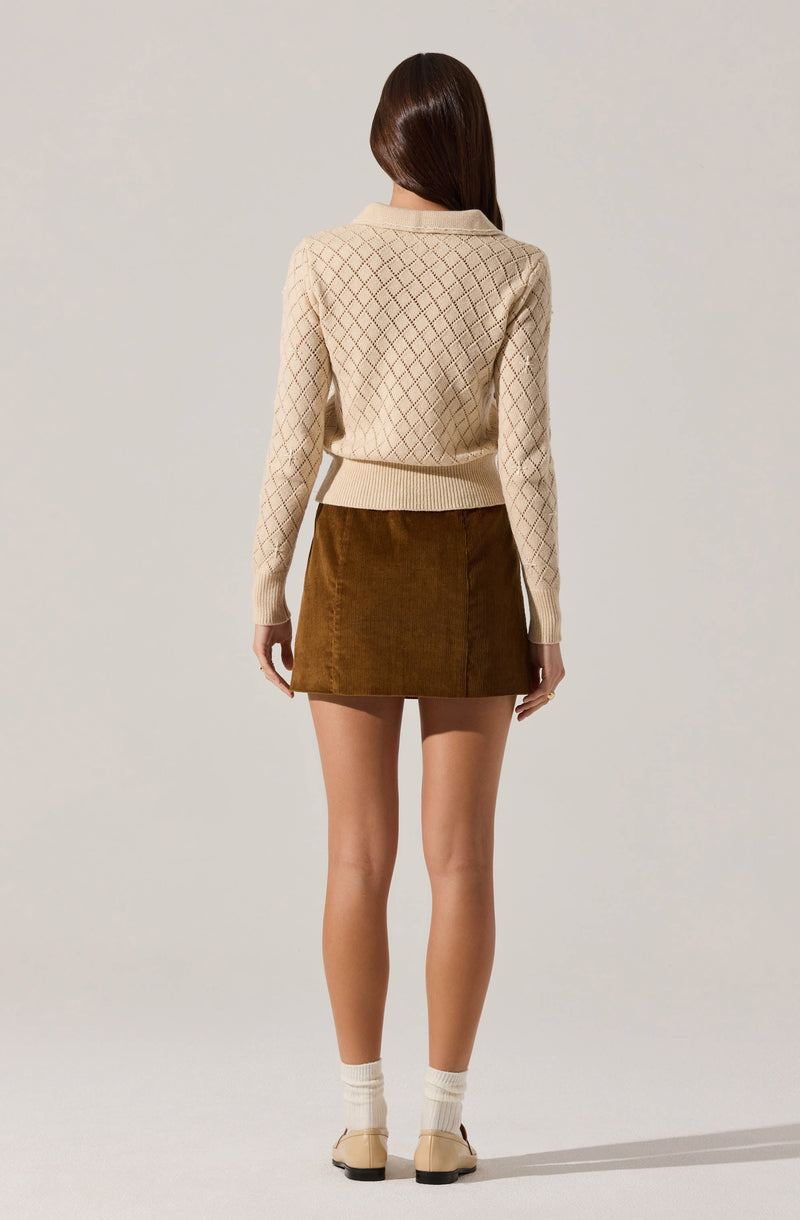 A person is wearing a cream-colored, long-sleeved sweater with a diamond-knit pattern, paired with a brown corduroy skirt. The individual is standing with their back to the camera, showing the sweater's fitted waistband and the skirt's above-the-knee length. They are also wearing beige shoes with low heels and cream-colored socks.
