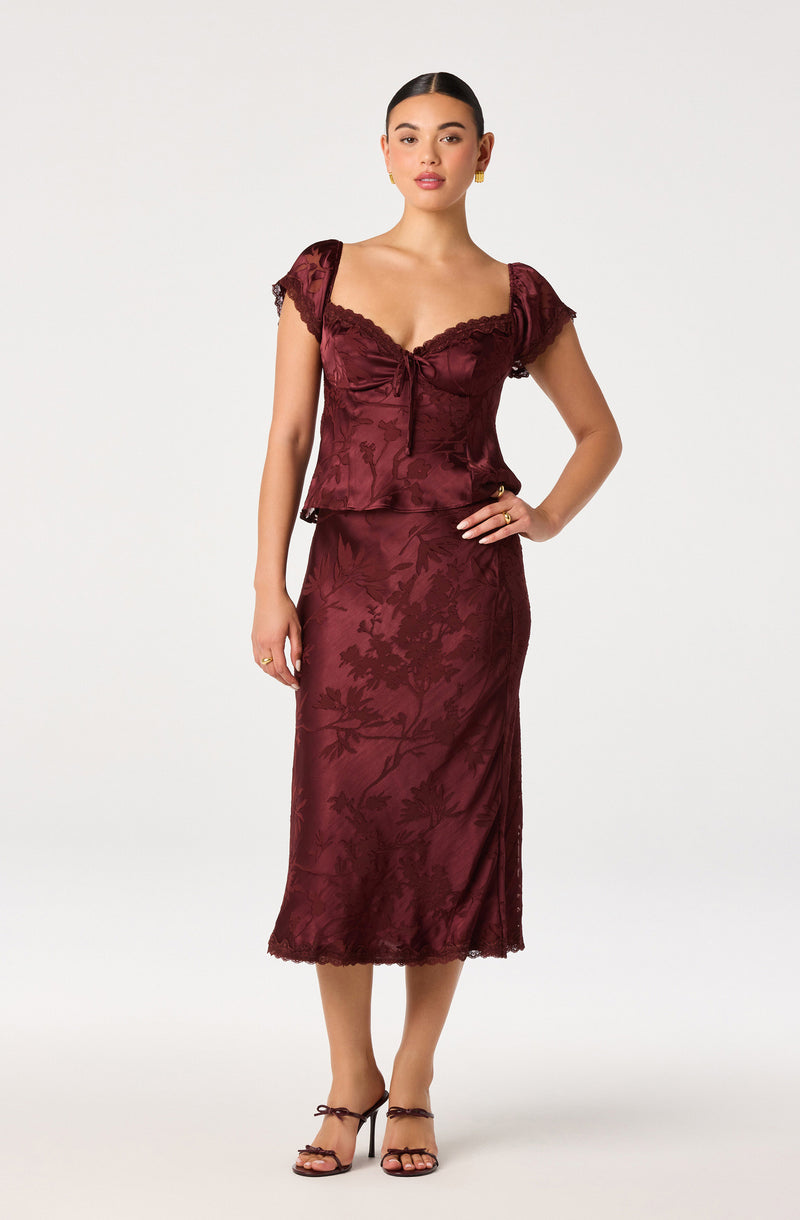 Woman wearing a burgundy satin matching top and skirt with floral embroidery, lace details, and tie straps on her heels.