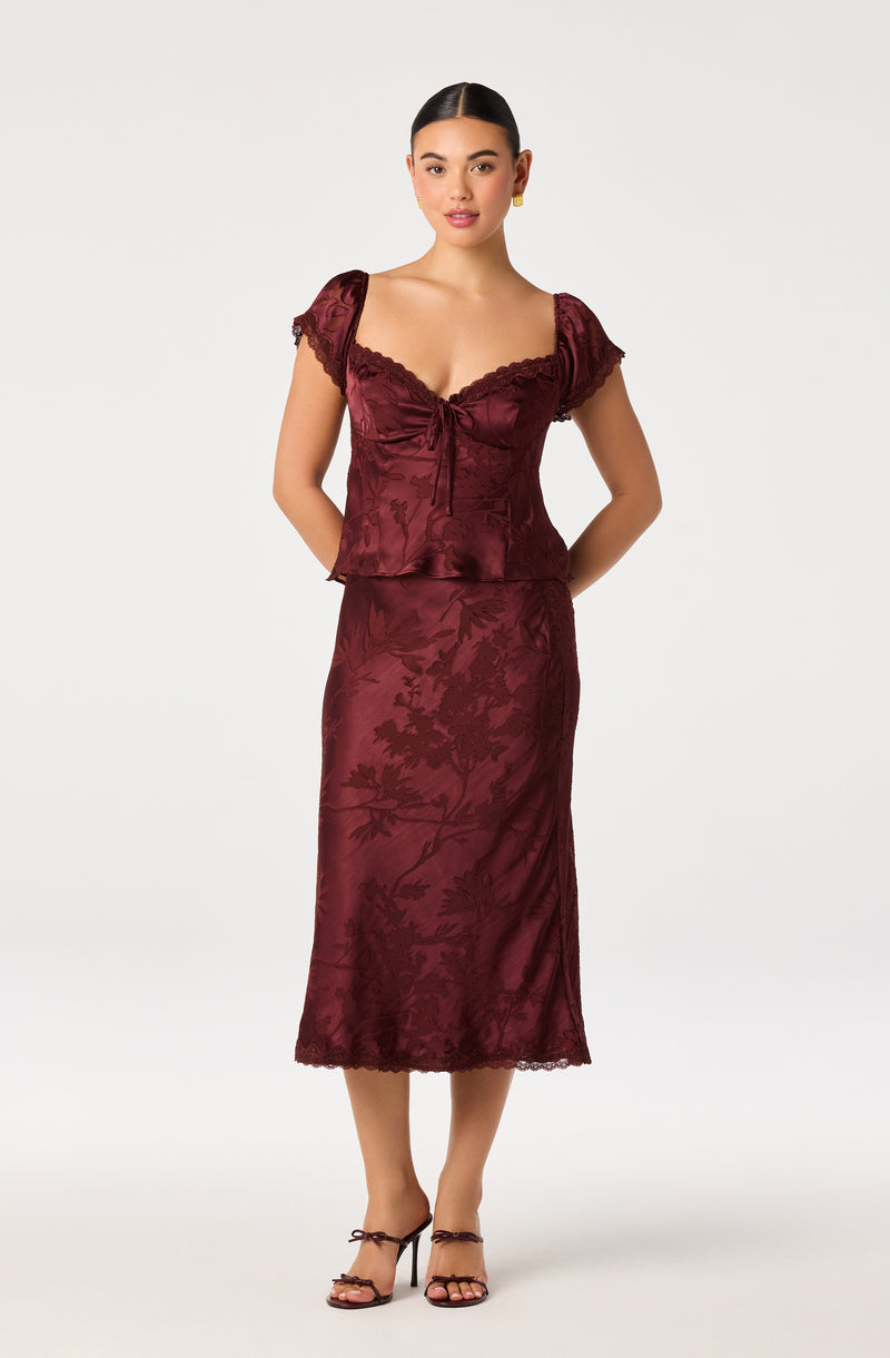 Woman wearing a burgundy satin two-piece dress with lace and floral patterns, and open-toe heels.