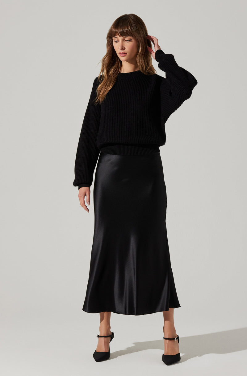 A woman is wearing a black, chunky knit sweater with long sleeves and a high neckline. She pairs it with a long, shiny black satin or leather maxi skirt that reaches her ankles. She completes the outfit with black high-heeled shoes featuring ankle straps.