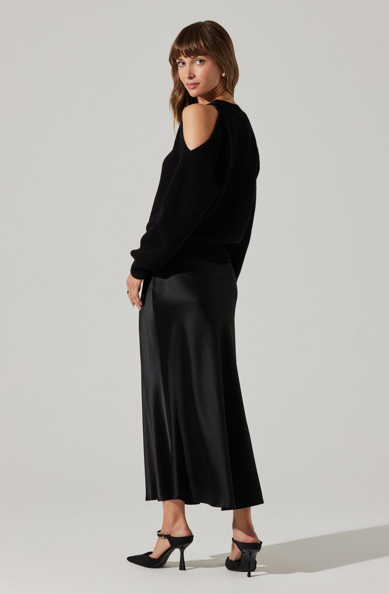 A woman is wearing a black sweater with shoulder cutouts and long sleeves. She pairs it with a long, black, satin-like skirt that reaches her ankles. She completes her outfit with black high-heeled shoes.
