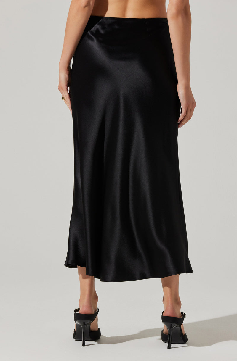 A woman is wearing a long, black satin skirt that reaches her ankles. The skirt has a smooth, shiny texture and a slightly flared silhouette. She is also wearing black high-heeled shoes with ankle straps.