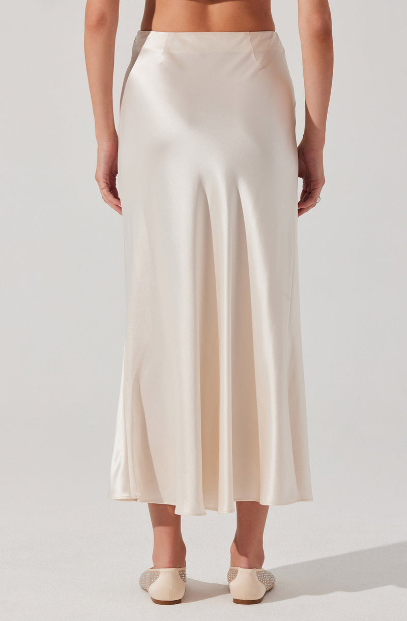 A woman is wearing a high-waisted, ankle-length cream-colored satin skirt with a smooth, shiny texture. The skirt has a relaxed fit with a slight flare at the bottom and features subtle pleats near the waistband. She is also wearing a pair of cream-colored shoes with decorative elements on the toes.