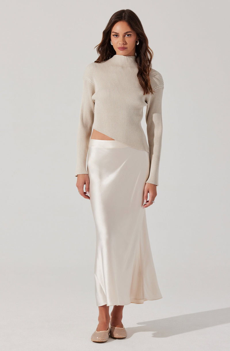 A woman is wearing a light beige, ribbed, long-sleeved sweater with a high neckline, which has a diagonal cutout on the side revealing a small part of her waist. She pairs it with a long, silky satin skirt in a matching cream or ivory color, that reaches her ankles. She completes the outfit with beige or nude-colored heels that have a textured pattern.