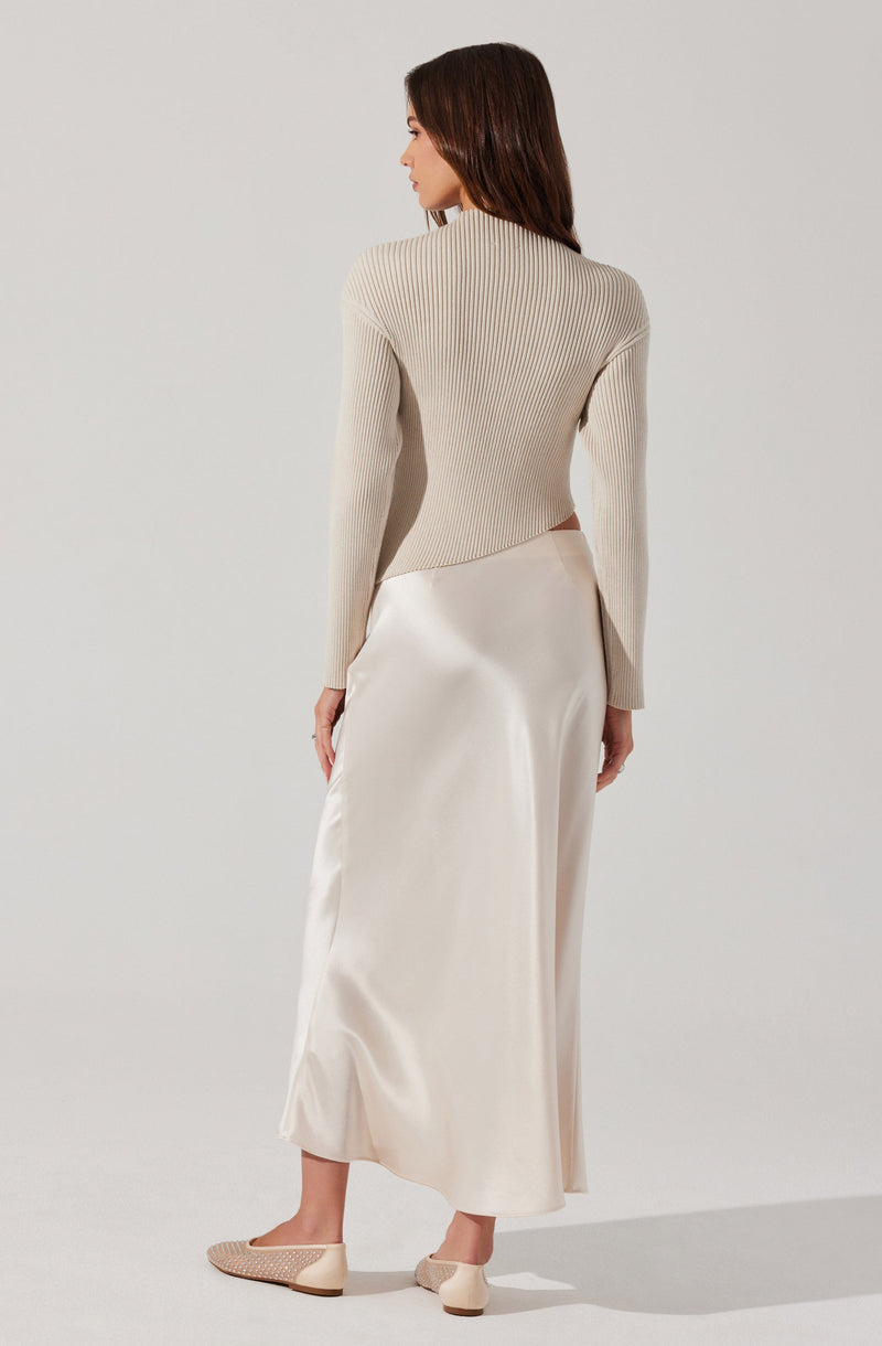 A woman is wearing a beige, long-sleeved, ribbed knit sweater with a high neckline. She is also dressed in an elegant, floor-length, satin-like skirt in a creamy off-white color. Her footwear consists of beige, low-heeled, woven-style shoes.