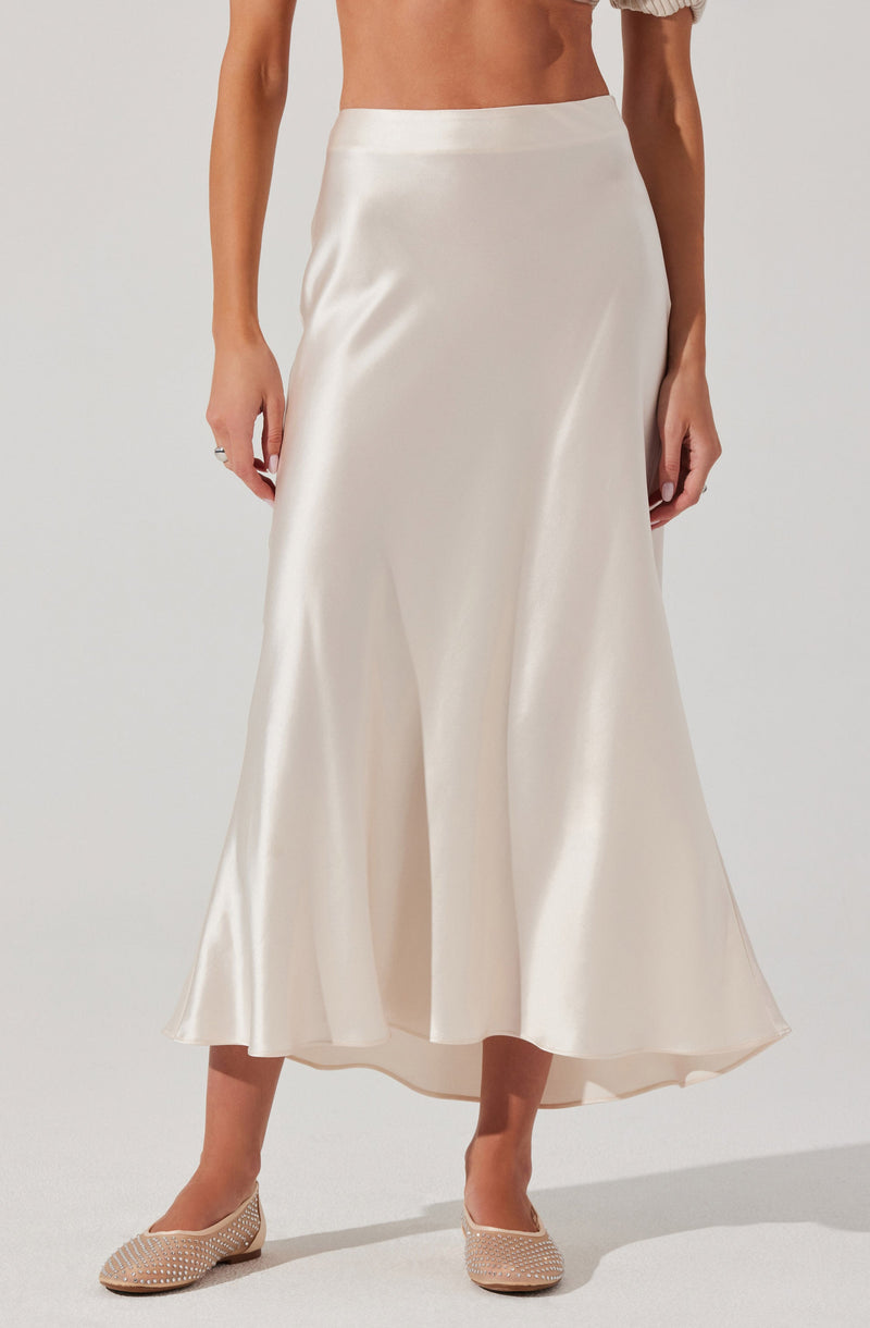 A woman is wearing a high-waisted, floor-length satin skirt in a shiny, ivory color. The skirt has a smooth, flowing fit with a slight flare at the hem. She is also wearing beige, pearl-embellished flats.