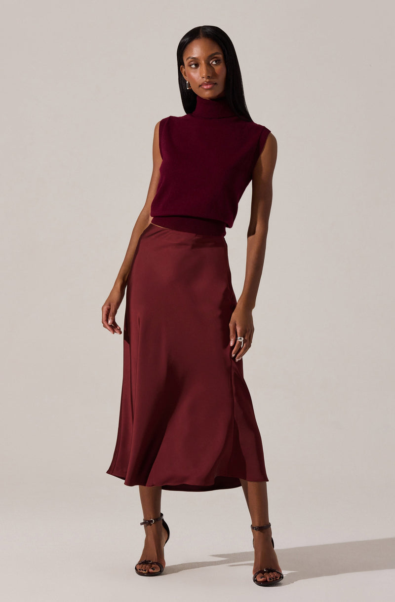 A woman is wearing a sleeveless, burgundy turtleneck top and a matching satin midi skirt with a slight slit on one side. She is also wearing black high-heeled sandals with ankle straps.