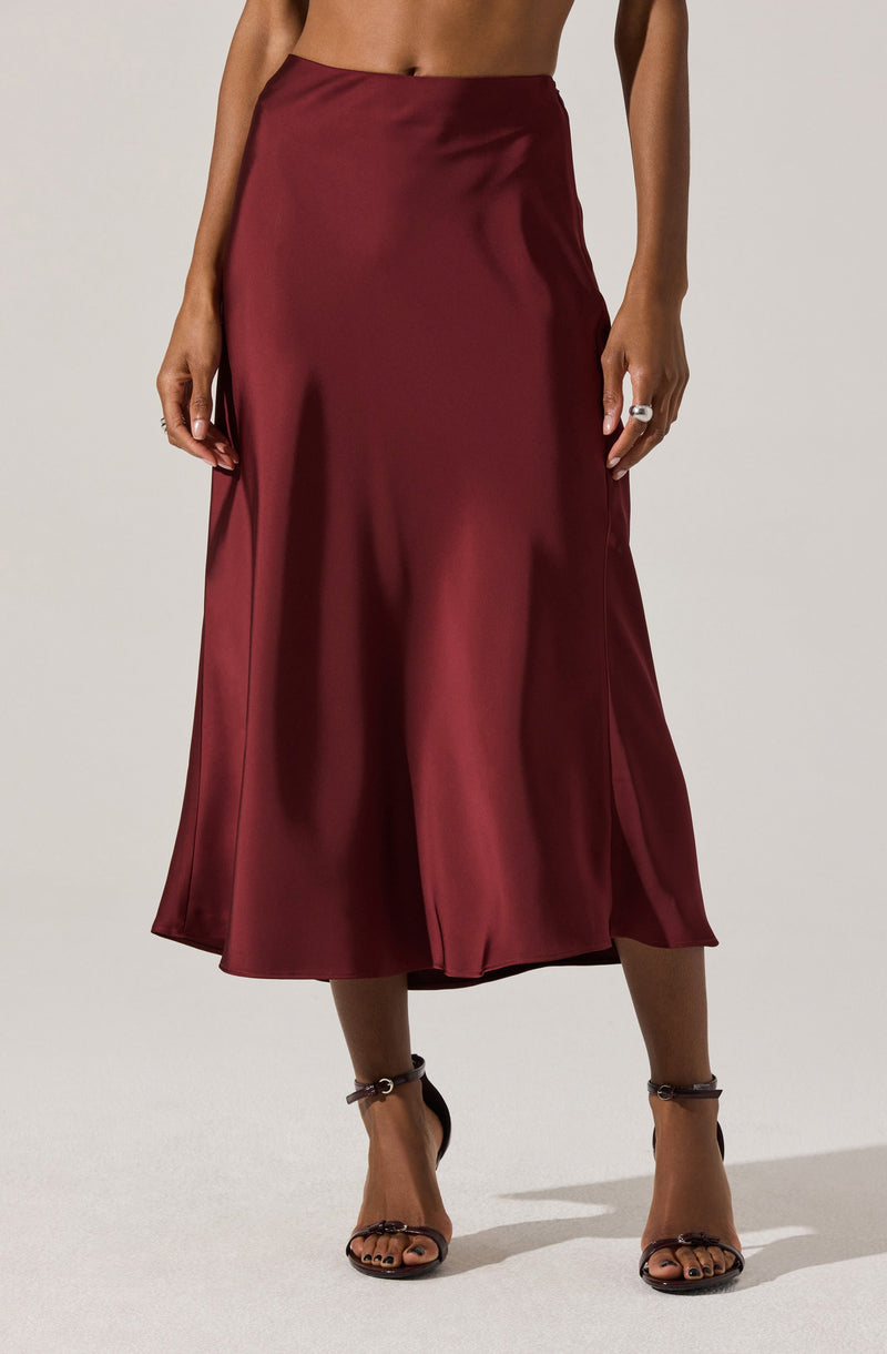 A woman is wearing a high-waisted, ankle-length satin skirt in a deep burgundy color. The skirt has a smooth, shiny texture and features a subtle slit on one side. She is also wearing black strappy high-heeled sandals that wrap around her ankles.